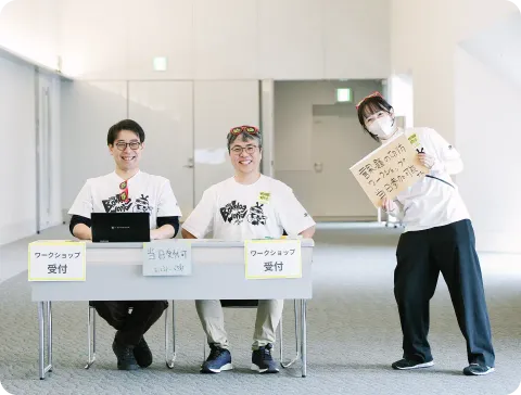 イベントのワークショップ受付デスク。お揃いの白いTシャツを着た3人のスタッフが、笑顔で参加者を迎えている。