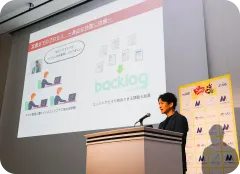 セミナー会場の壇上で話す男性。背後のスクリーンには「Backlog」を活用した業務プロセスの定着についての図解が投影されている。