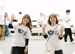 イベント会場で、お揃いのイベントTシャツを着てカメラに向かって手を振る、笑顔の女性スタッフ2人。