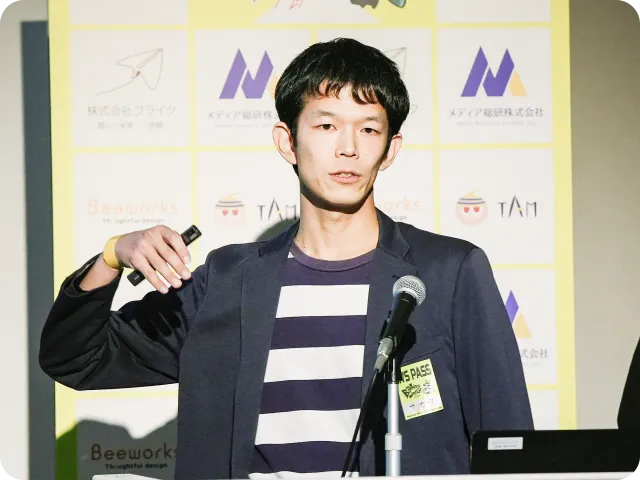 企業ロゴが並ぶパネルの前で、マイクを持ちプレゼンテーションを行うジャケット姿の男性。