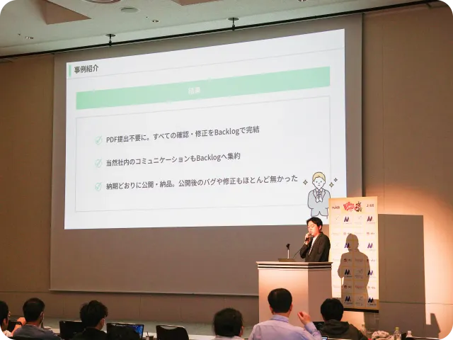 講演会場で、大きなスクリーンに投影されたスライドを使ってプレゼンテーションが行われている様子。